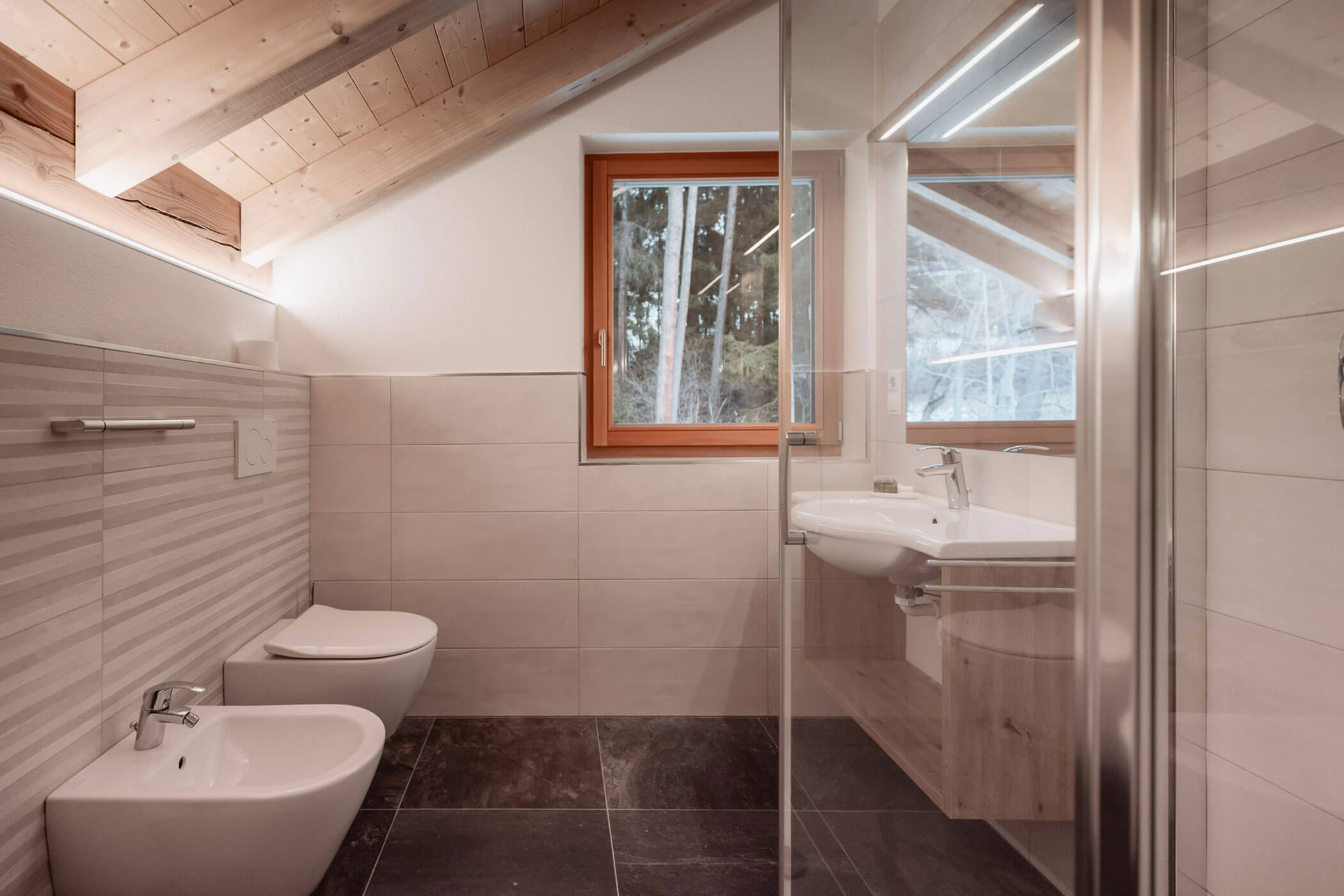 Modernes Badezimmer mit schräger Holzdecke, gefliesten Wänden, einem Bidet, einer Toilette, einem Waschbecken und einer Duschkabine aus Glas. - Lantl Deluxe Apartments