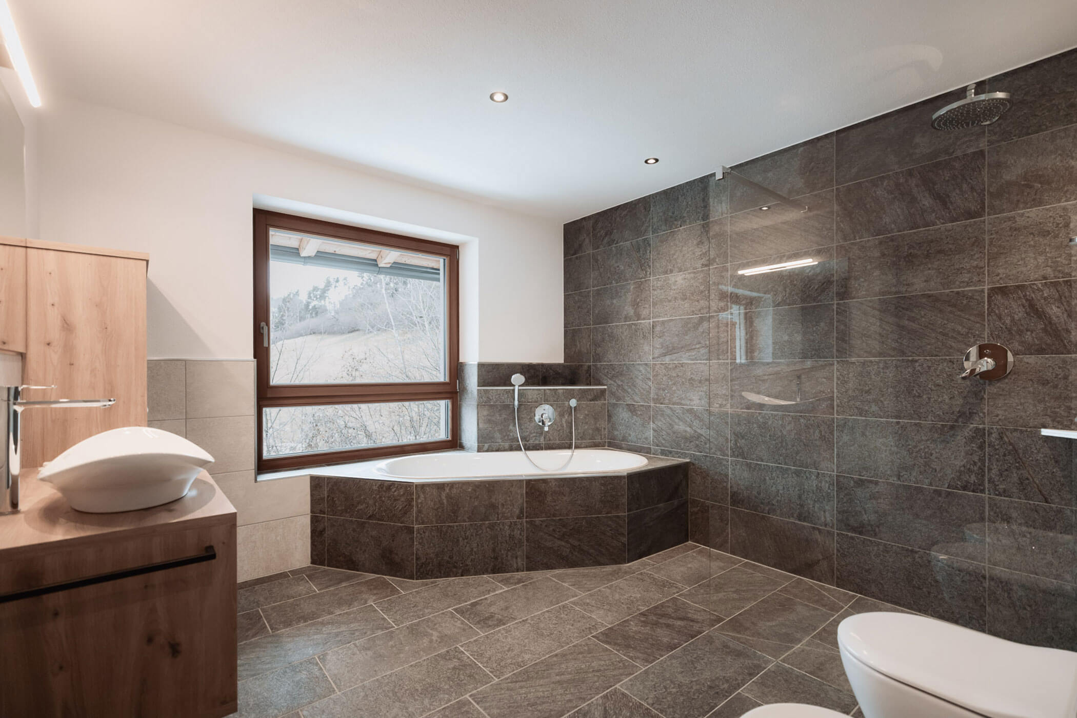 Modernes Badezimmer mit großflächig gefliesten Wänden, einer Badewanne am Fenster und einem Aufsatzwaschbecken auf einem Waschtisch aus Holz. - Lantl Deluxe Apartments