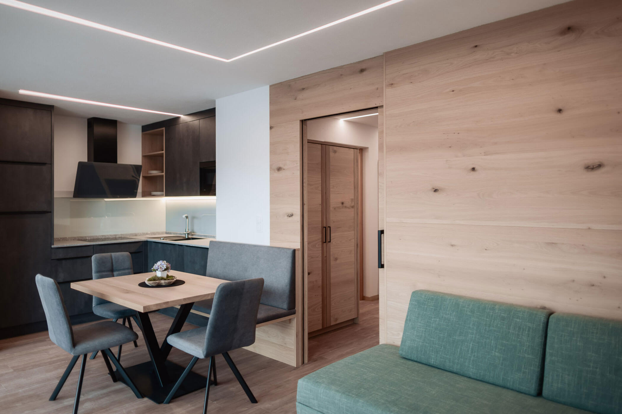 Moderne Wohnung mit einem Esstisch aus Holz, grauen Stühlen, einem grünen Sofa und einer minimalistischen Küche. - Lantl Deluxe Apartments
