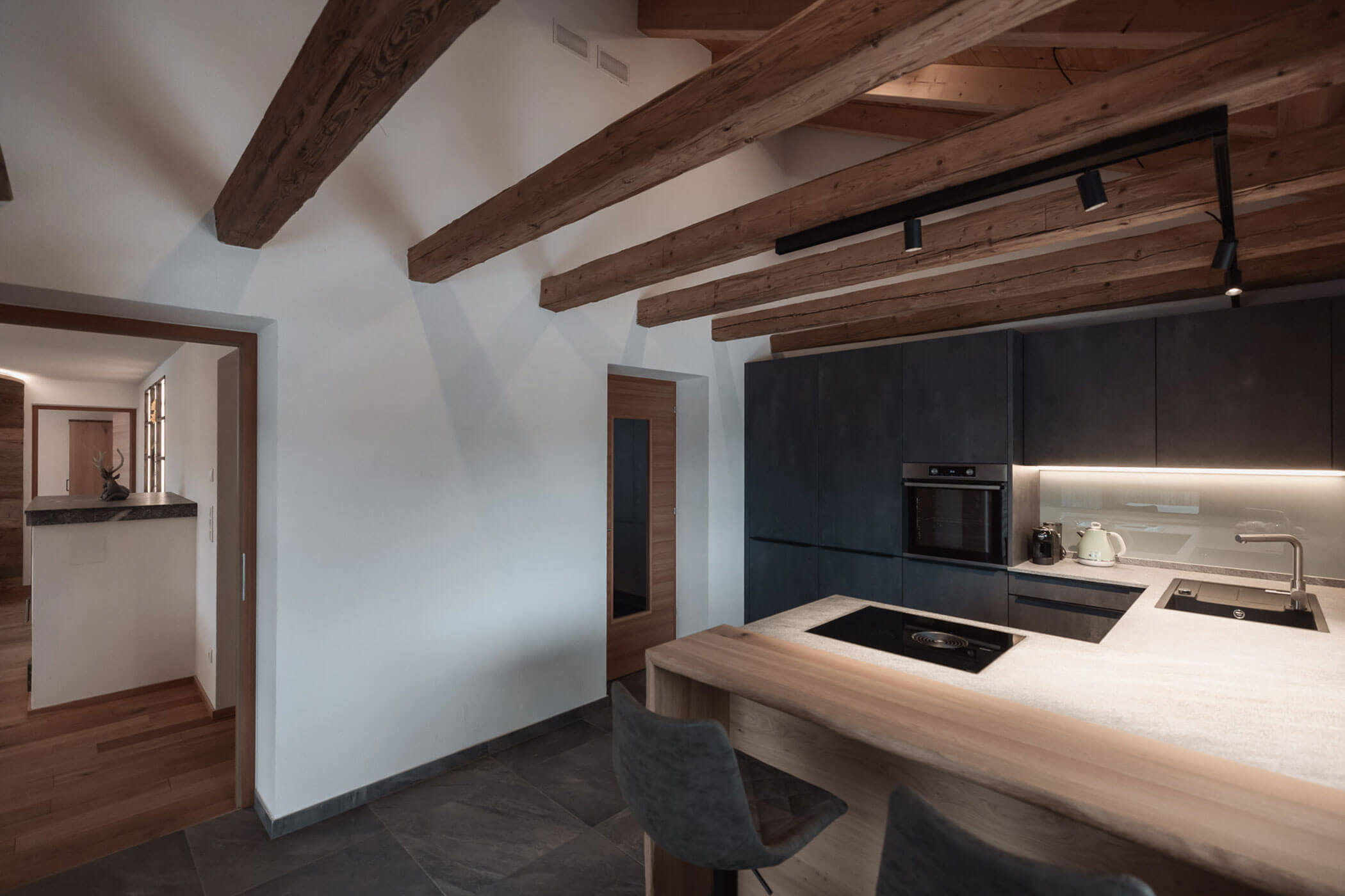 Moderne Küche mit dunklen Schränken, freiliegenden Holzbalken und einer Frühstückstheke aus Holz mit zwei Stühlen. - Lantl Deluxe Apartments