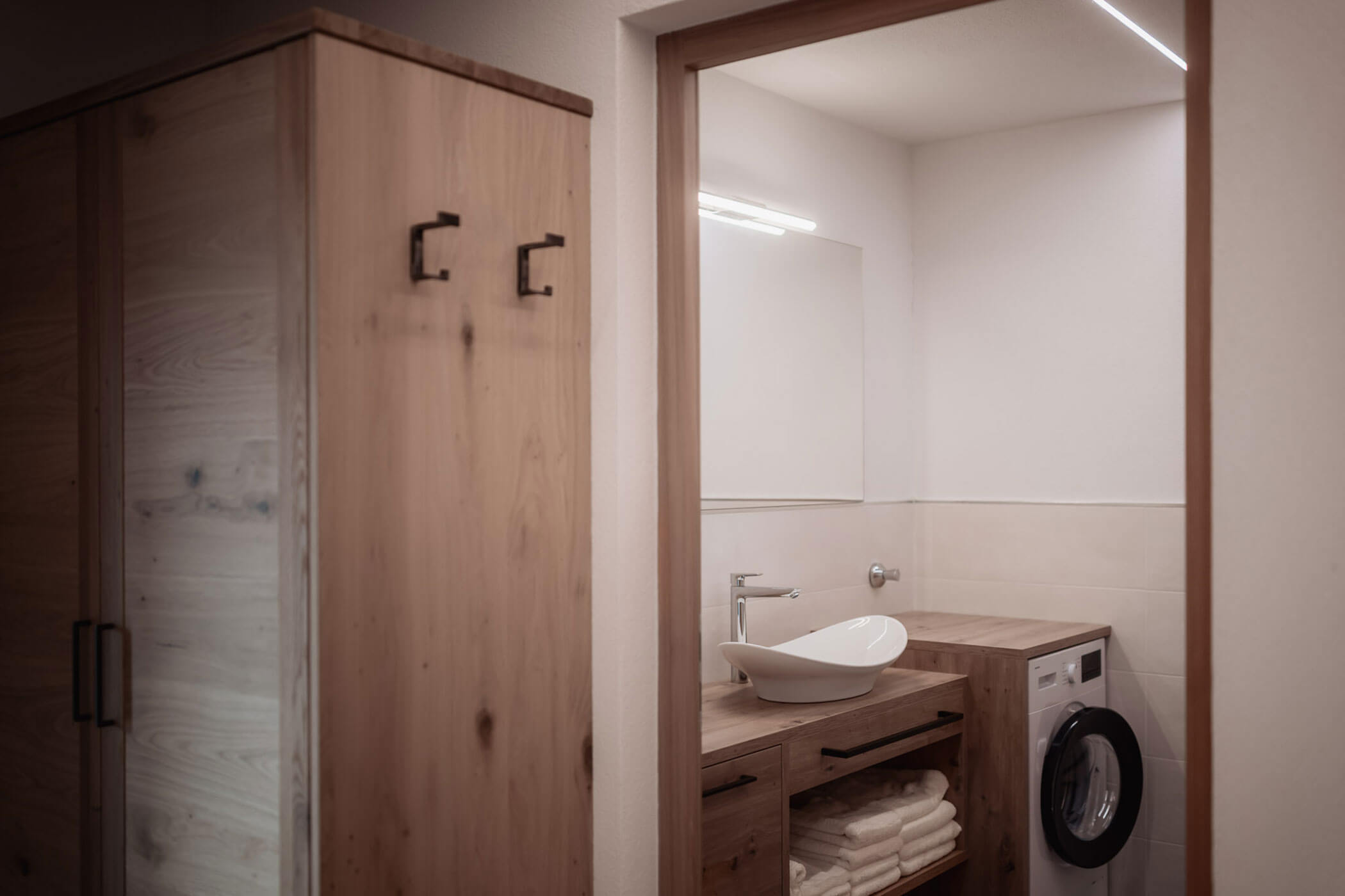 Kleines Badezimmer mit Holzschränken, einem Aufsatzwaschbecken, gestapelten Handtüchern und einer Waschmaschine unter dem Waschtisch. - Lantl Deluxe Apartments