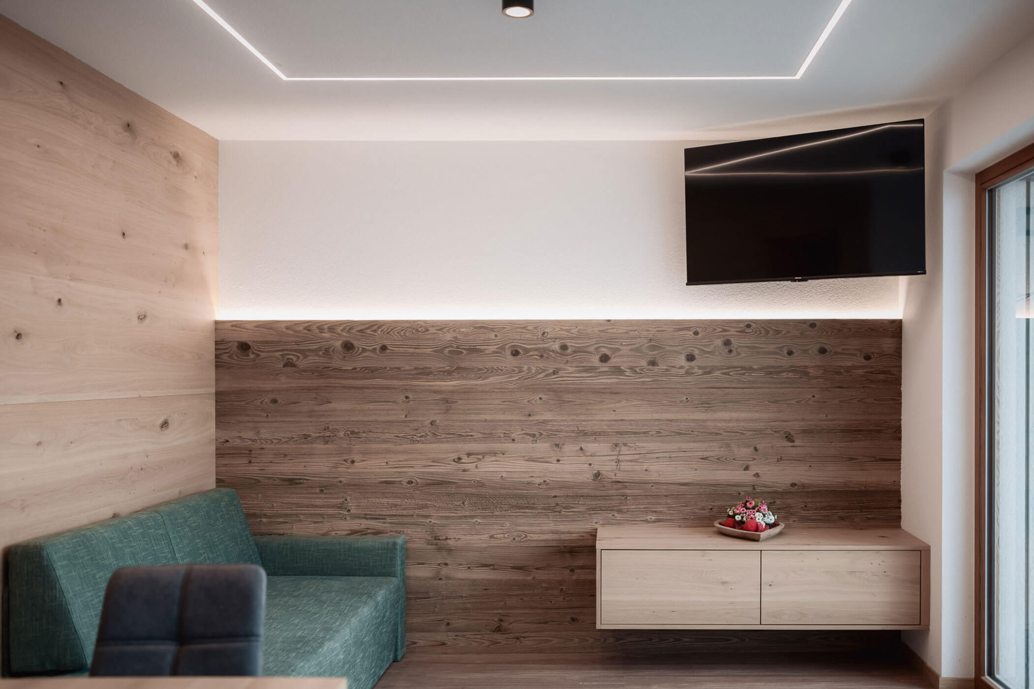 Minimalistisches Wohnzimmer mit Holzwänden, einem grünen Sofa, einem an der Wand montierten Fernseher und einem niedrigen Schrank mit einer Obstschale. - Lantl Deluxe Apartments