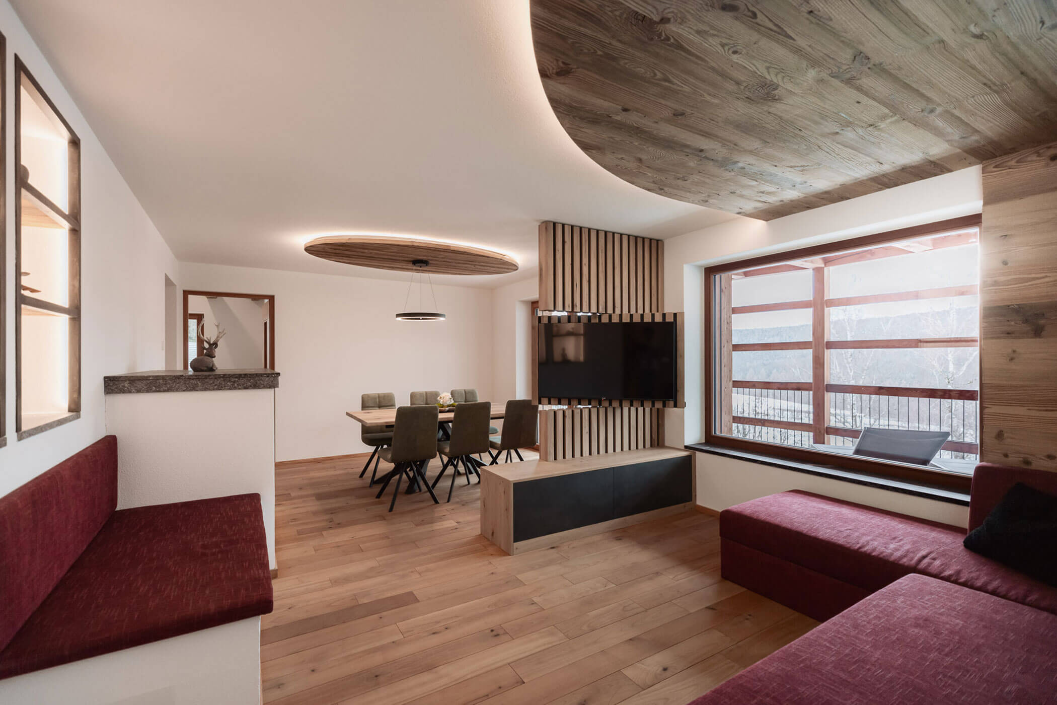 Moderner Wohn- und Essbereich mit Holzböden, roten Sitzmöbeln und großen Fenstern, durch die viel Tageslicht hereinströmt. - Lantl Deluxe Apartments