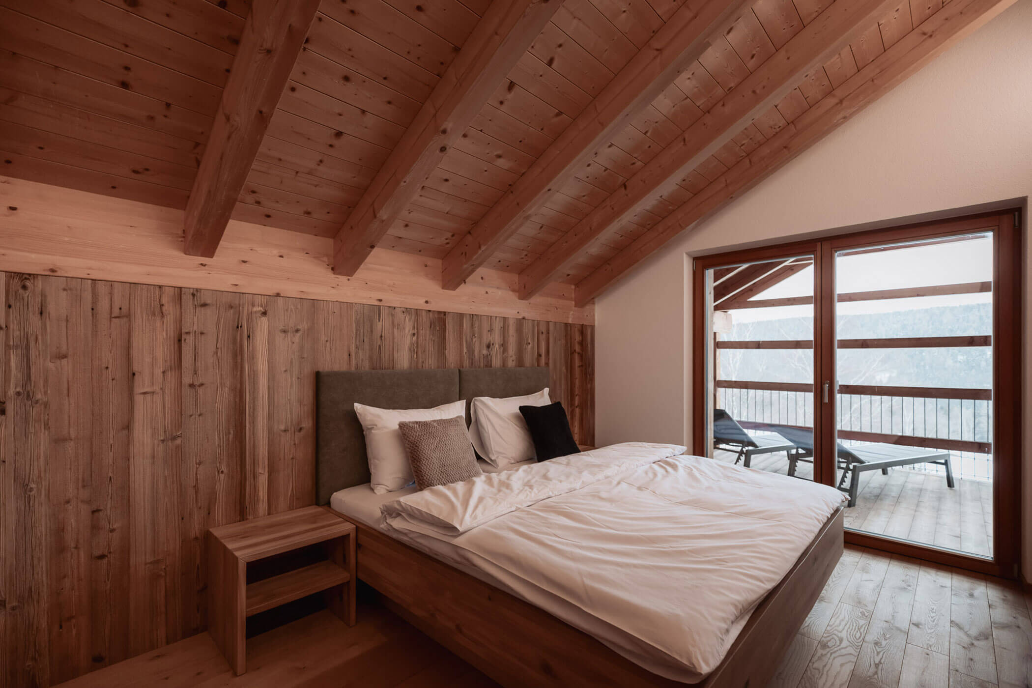 Gemütliches Schlafzimmer im Landhausstil mit Holzwänden und -decke, einem großen Bett und Glastüren, die auf einen Balkon mit Liegestühlen führen. - Lantl Deluxe Apartments