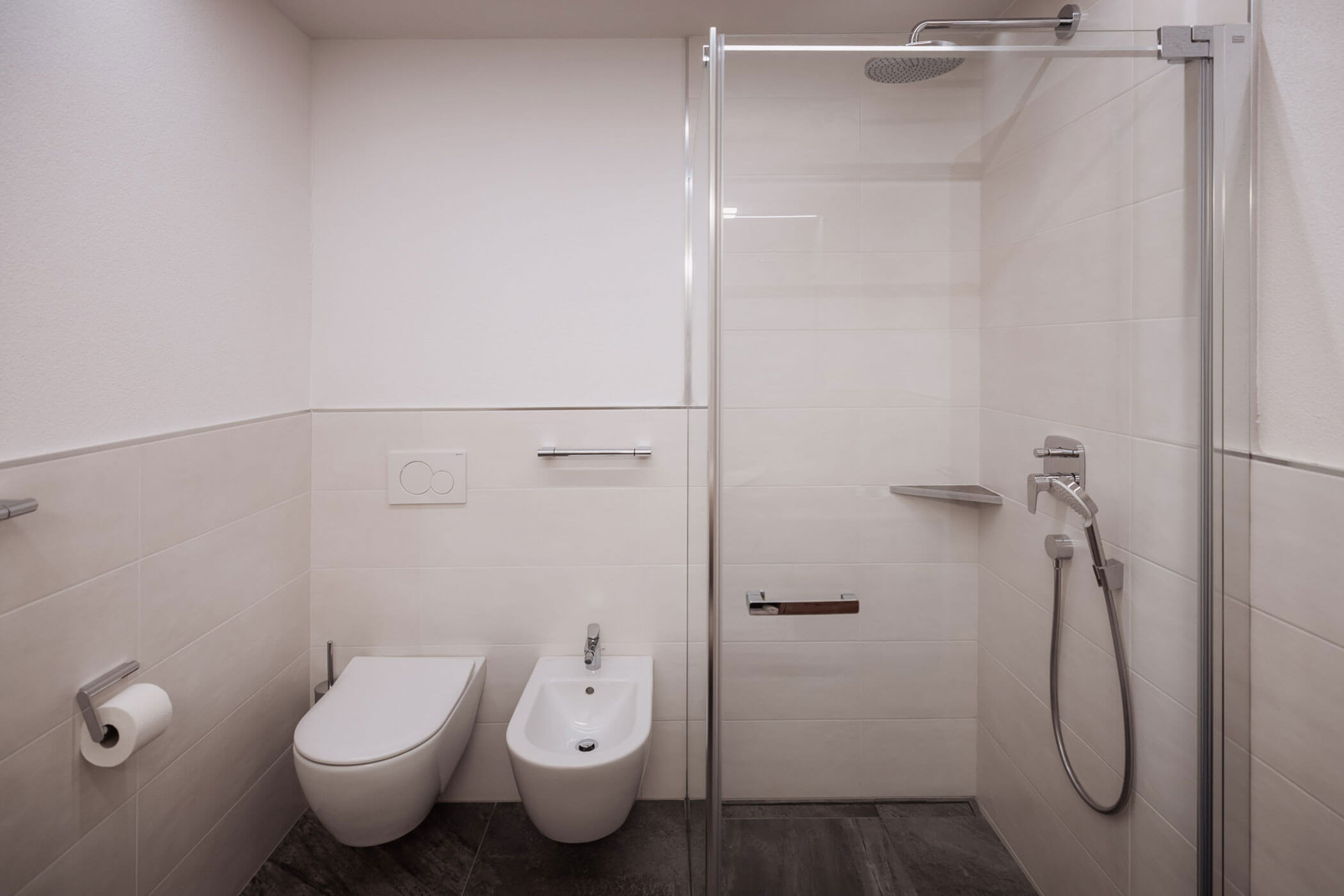 Modernes Badezimmer mit wandhängendem WC, Bidet, Duschkabine aus Glas und beigen Fliesenwänden. - Lantl Deluxe Apartments