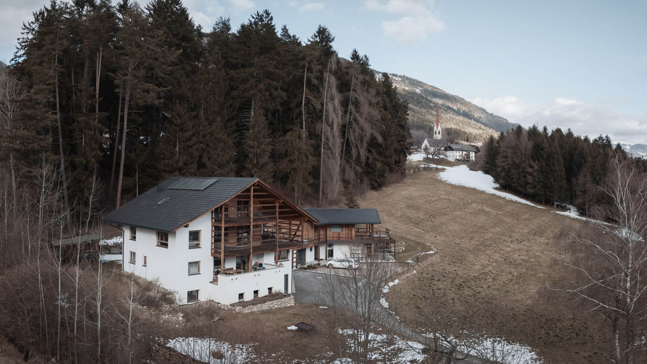 Ein weißes Haus mit Holzbalkonen neben einem bewaldeten Hügel, mit Schneeflecken und einer Kirche in der Ferne. - Lantl Deluxe Apartments
