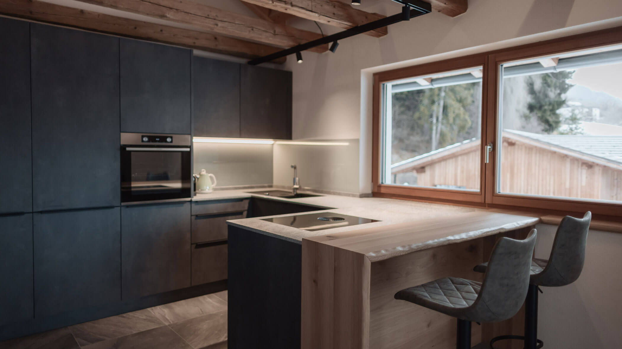 Moderne Küche mit dunklen Schränken, Holzakzenten, einer Bar mit Sitzgelegenheiten und einem großen Fenster mit malerischem Blick ins Freie. - Lantl Deluxe Apartments