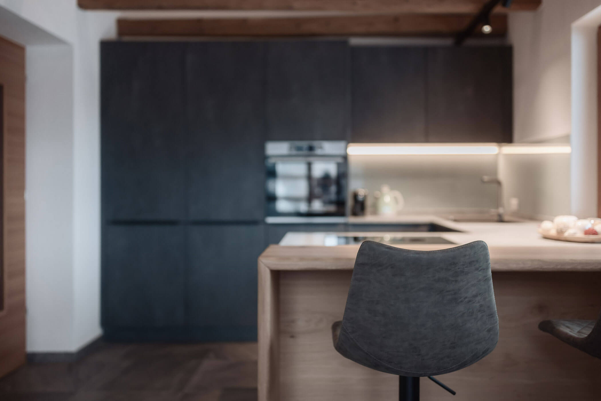 Moderne Küche mit dunklen Schränken, einem Einbaubackofen und einem grauen Barhocker an einer Holztheke. - Lantl Deluxe Apartments
