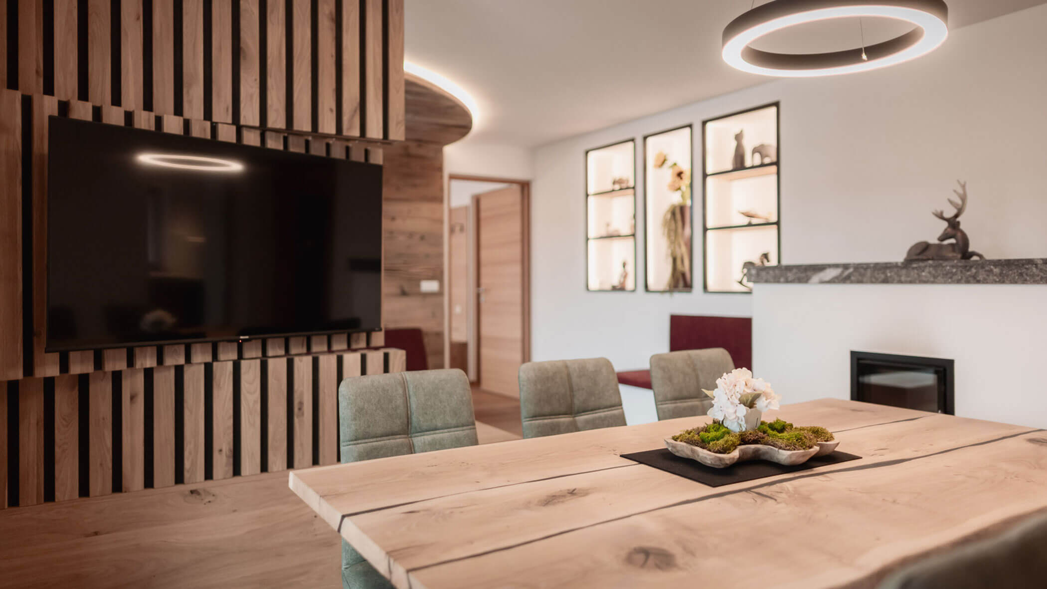 Moderner Ess- und Wohnbereich mit Holzakzenten, einem großen Fernseher sowie dekorativen Wandregalen und Leuchten. - Lantl Deluxe Apartments