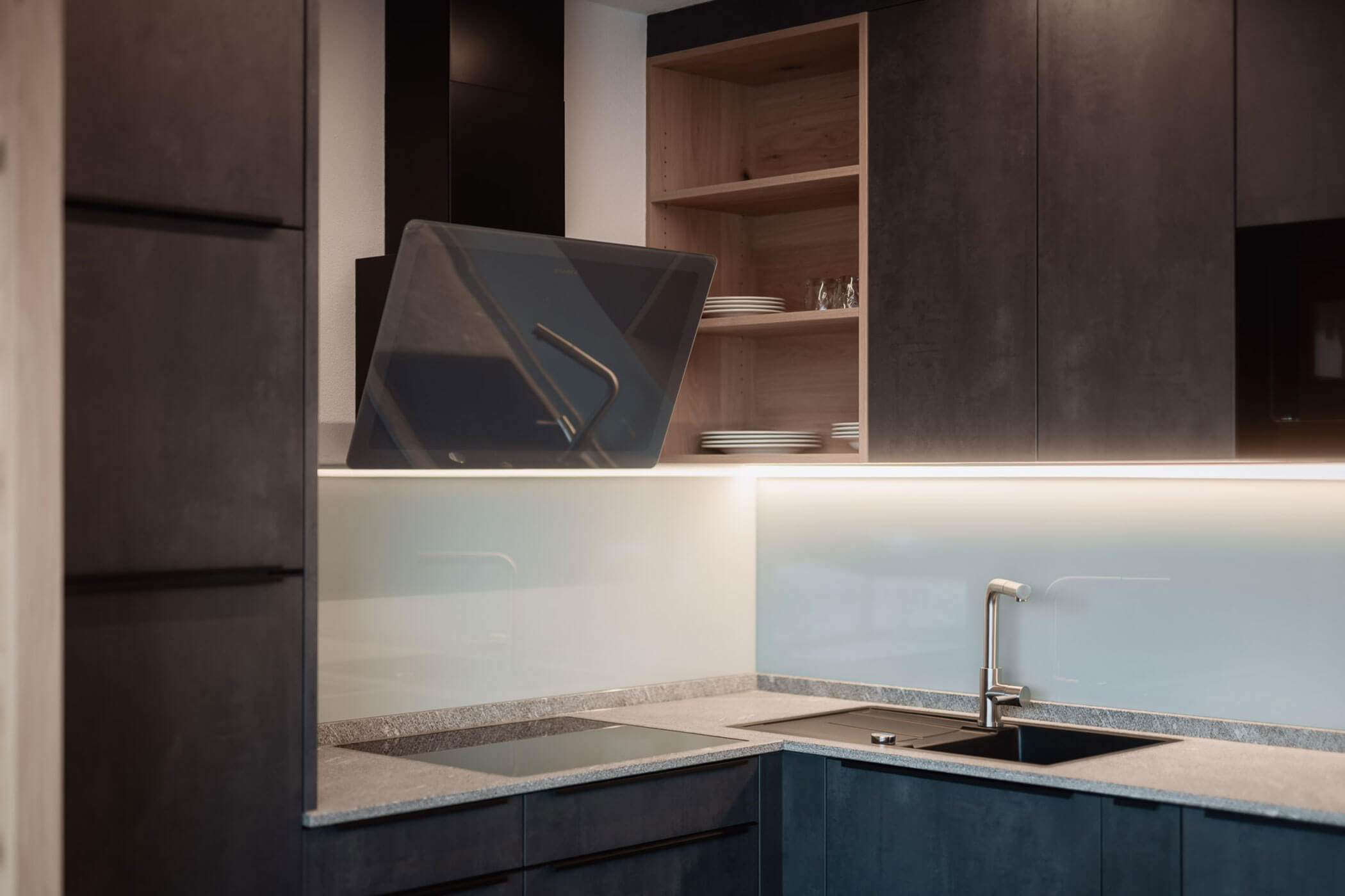 Moderne Küche mit dunklen Schränken, offenen Regalen mit Tellern, einer Glas-Küchenrückwand und einer Unterbauleuchte. - Lantl Deluxe Apartments