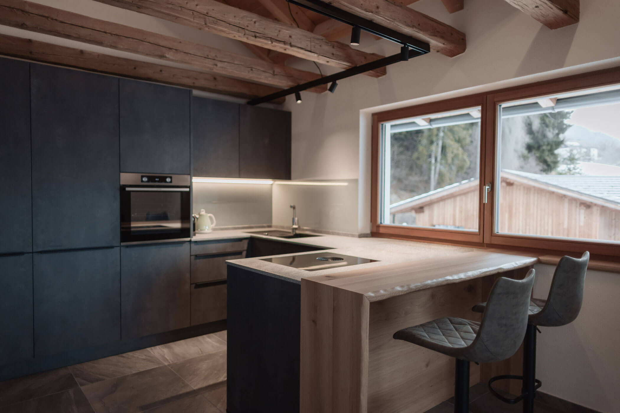 Moderne Küche mit dunklen Schränken, Holzakzenten und zwei Barhockern am Fenster mit Blick auf die Berge. - Lantl Deluxe Apartments