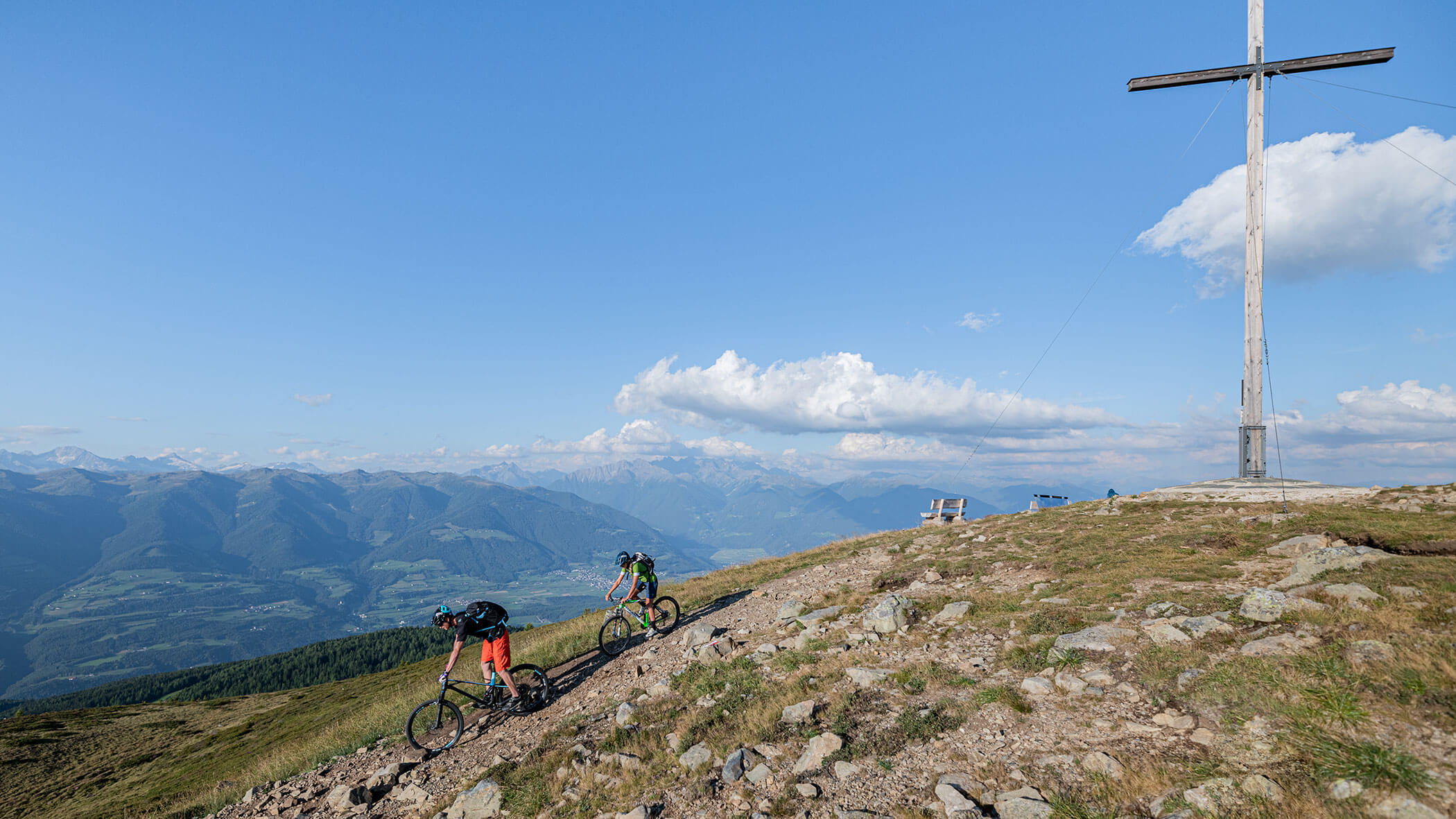 Zwei Radfahrer fahren auf einem Bergweg in der Nähe eines großen Kreuzes bergauf, im Hintergrund sind malerische Berge zu sehen. - Lantl Deluxe Apartments