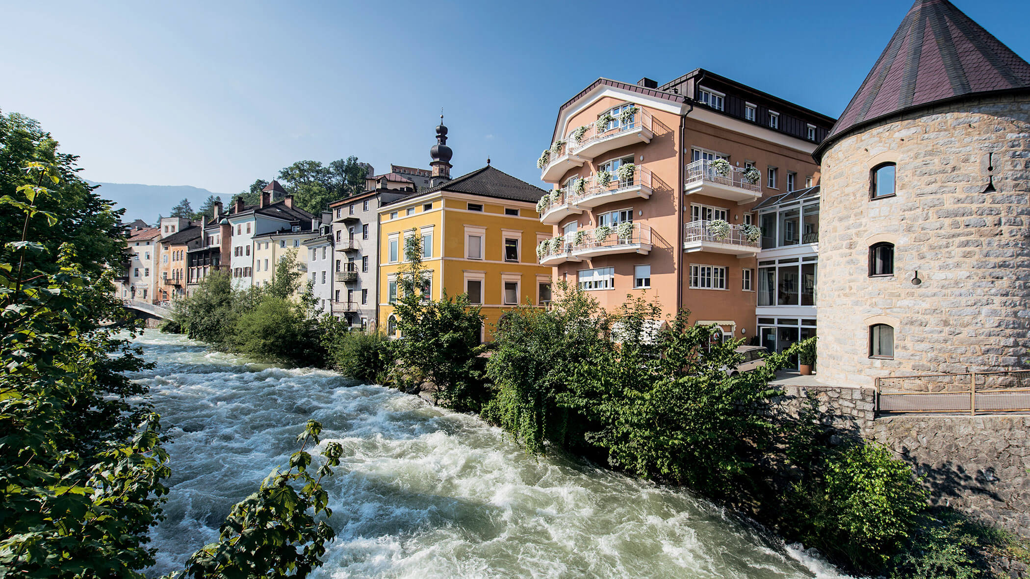 Farbenfrohe Gebäude mit Balkonen am Ufer eines reißenden Flusses unter strahlend blauem Himmel. - Lantl Deluxe Apartments