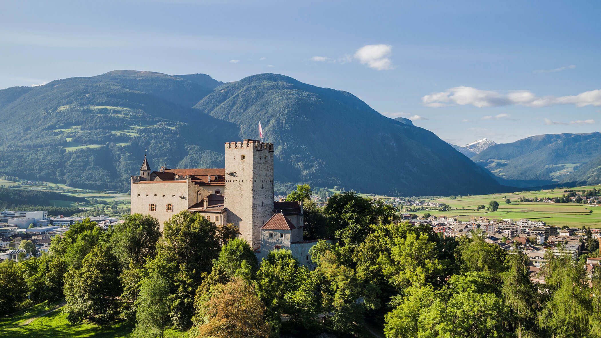 Mittelalterliche Burg auf einem Hügel, umgeben von Bäumen, mit Bergen und einer kleinen Stadt im Hintergrund. - Lantl Deluxe Apartments