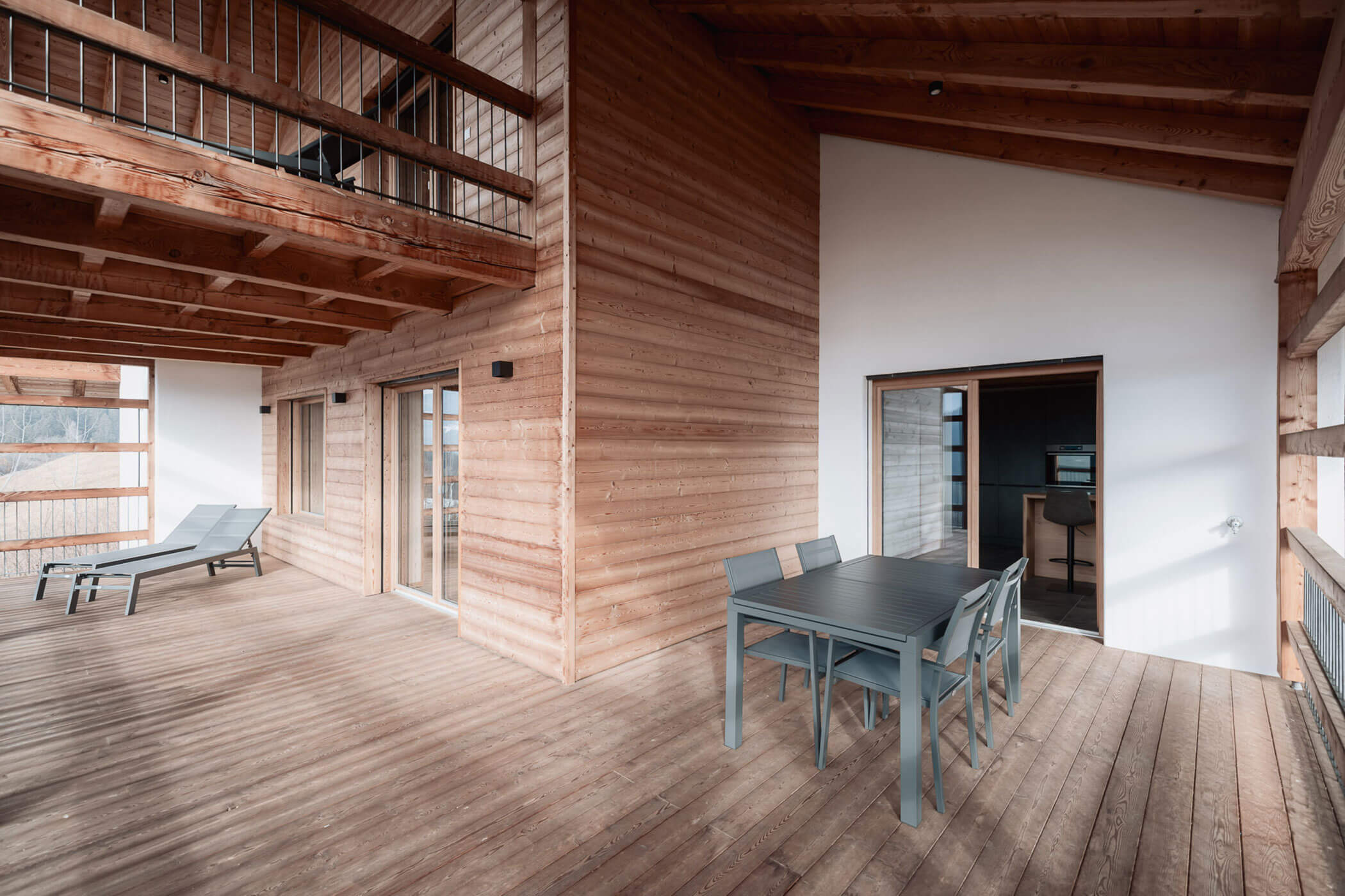 Moderne Holzterrasse mit Esstisch, Stühlen und Liegestühlen, umgeben von Holzvertäfelungen und Geländern. - Lantl Deluxe Apartments