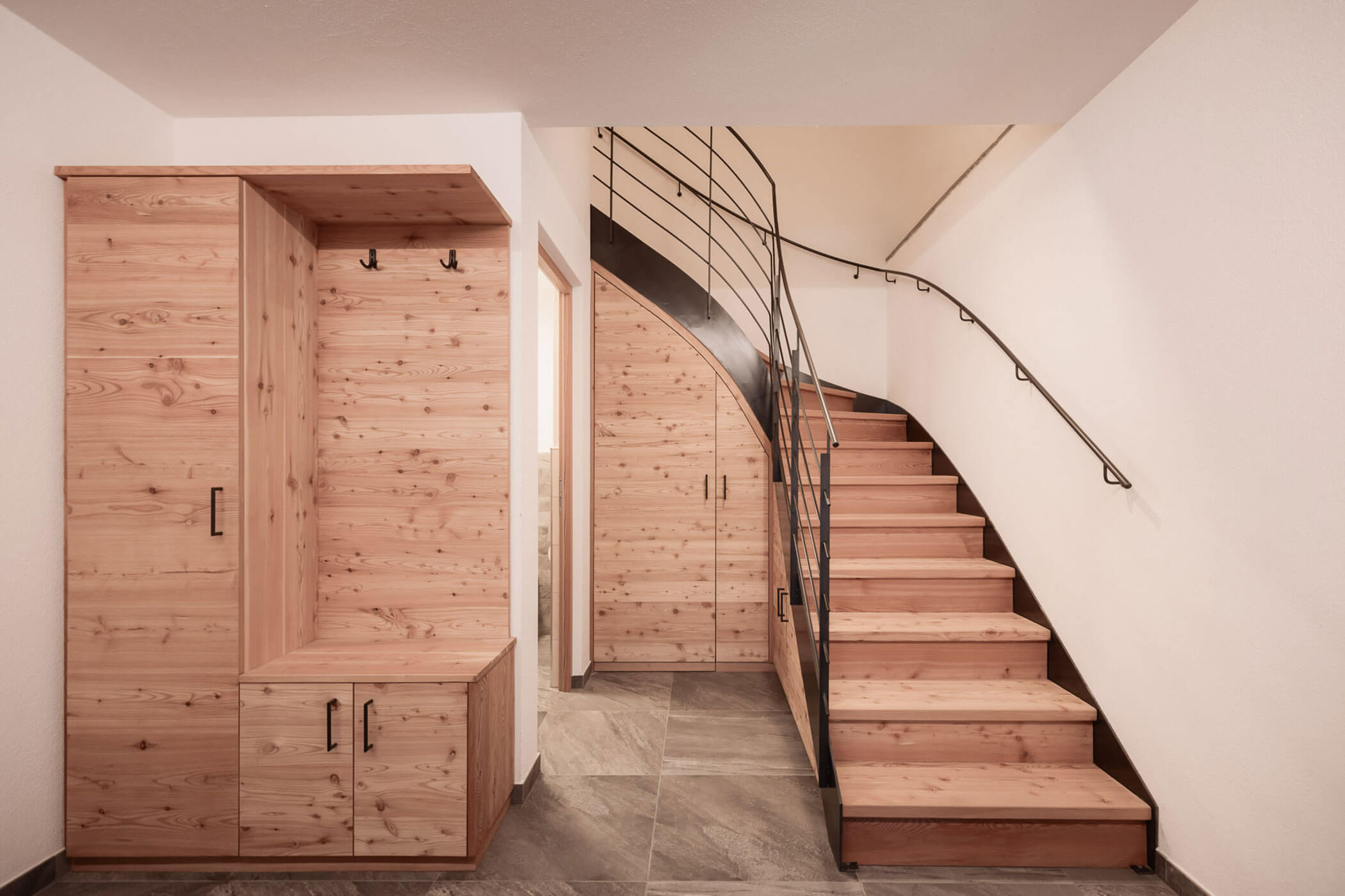 Moderner Flur mit Aufbewahrungsschränken aus Holz, Garderobenhaken und einer Treppe mit schwarzem Metallgeländer. - Lantl Deluxe Apartments