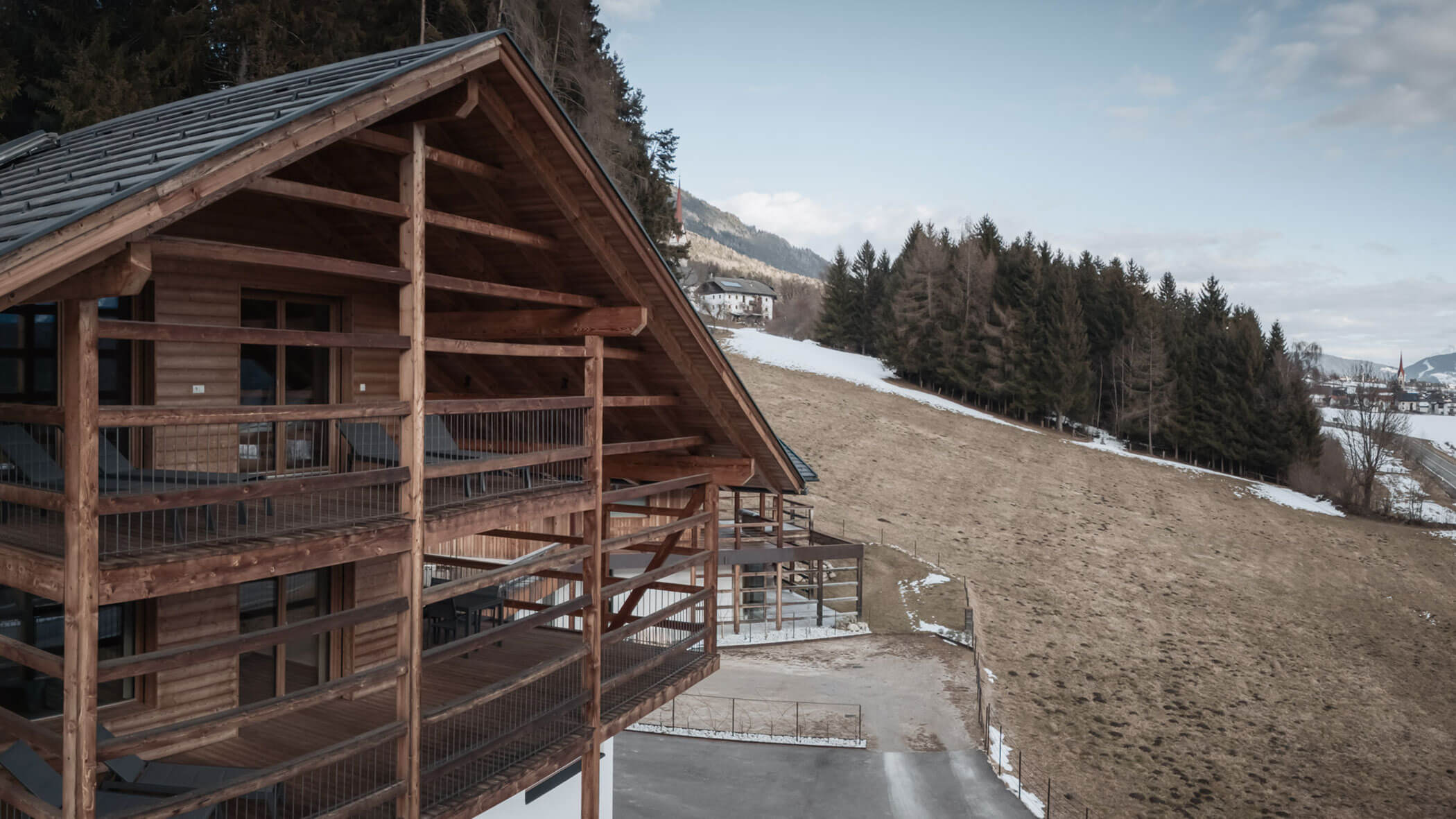 Holzchalet mit Balkonen an einem Hang, teilweise verschneite Landschaft und Kiefern im Hintergrund. - Lantl Deluxe Apartments