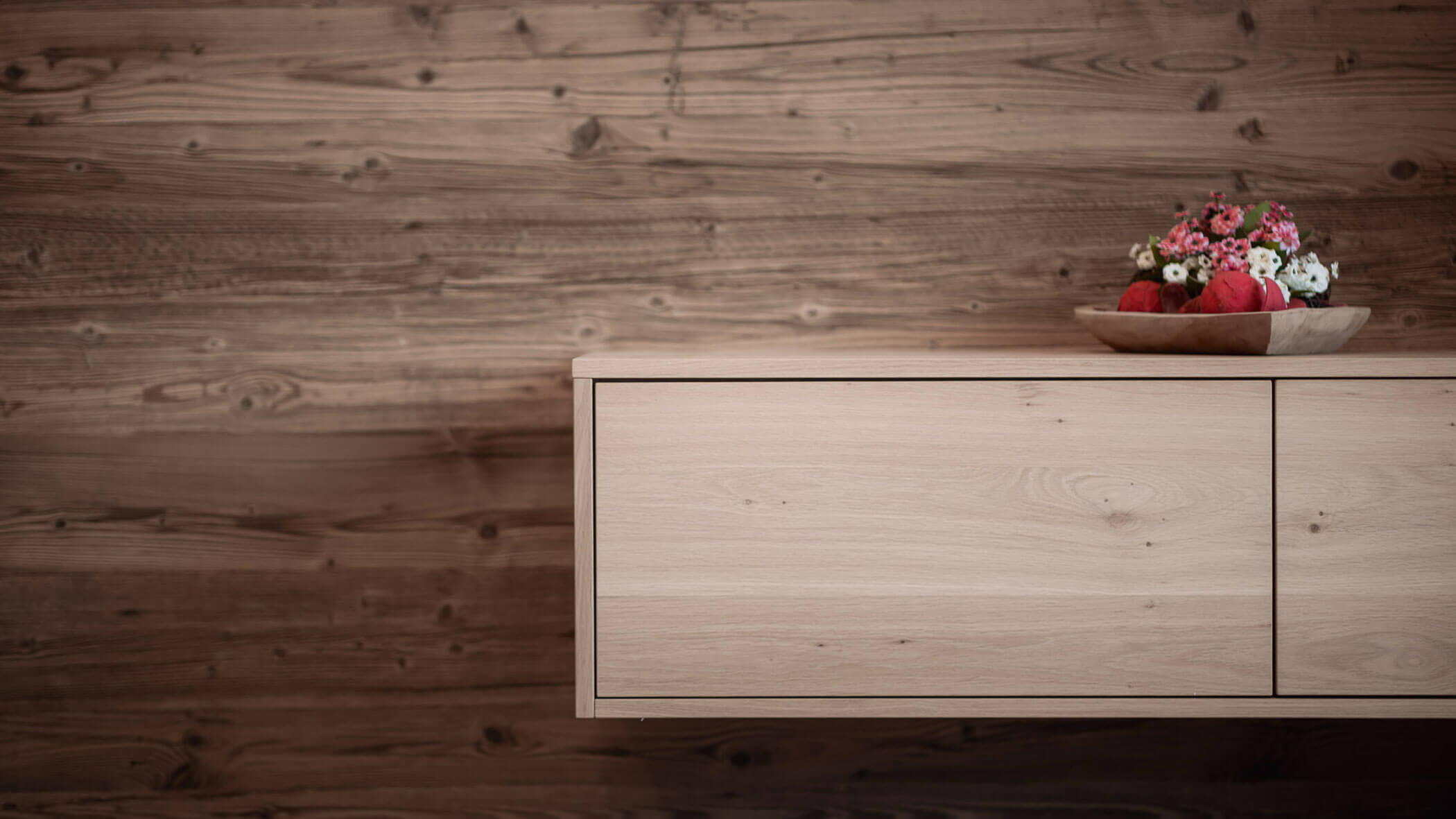 Ein schlichter Holzschrank, auf dem eine Schale mit bunten Blumen steht, vor dem Hintergrund einer Holzwand. - Lantl Deluxe Apartments