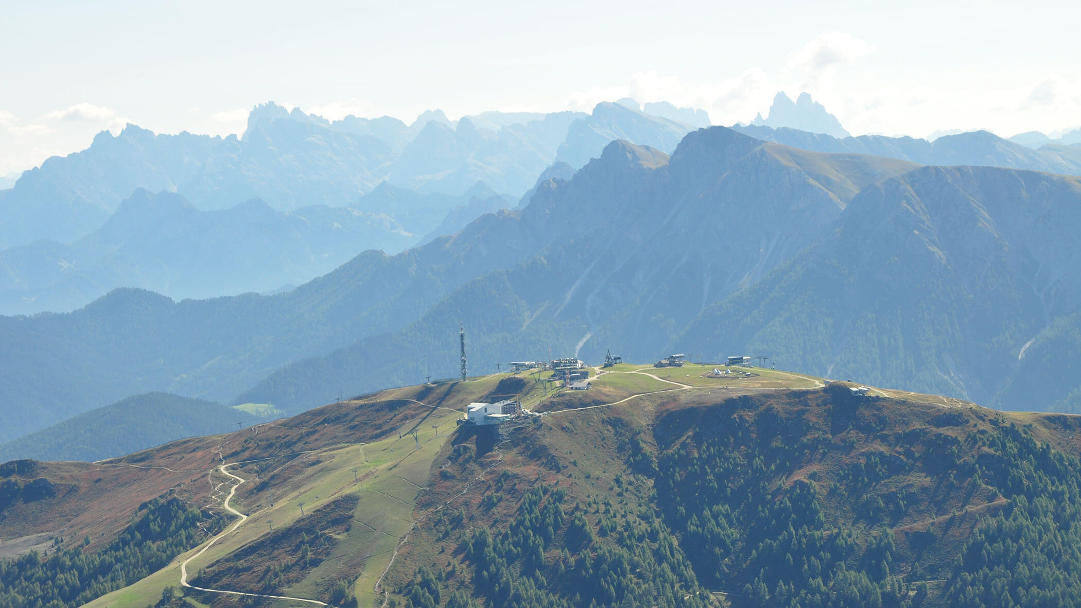 Berglandschaft mit Hügeln, kurvenreichen Straßen, Gebäuden und fernen Gipfeln unter einem klaren Himmel. - Lantl Deluxe Apartments