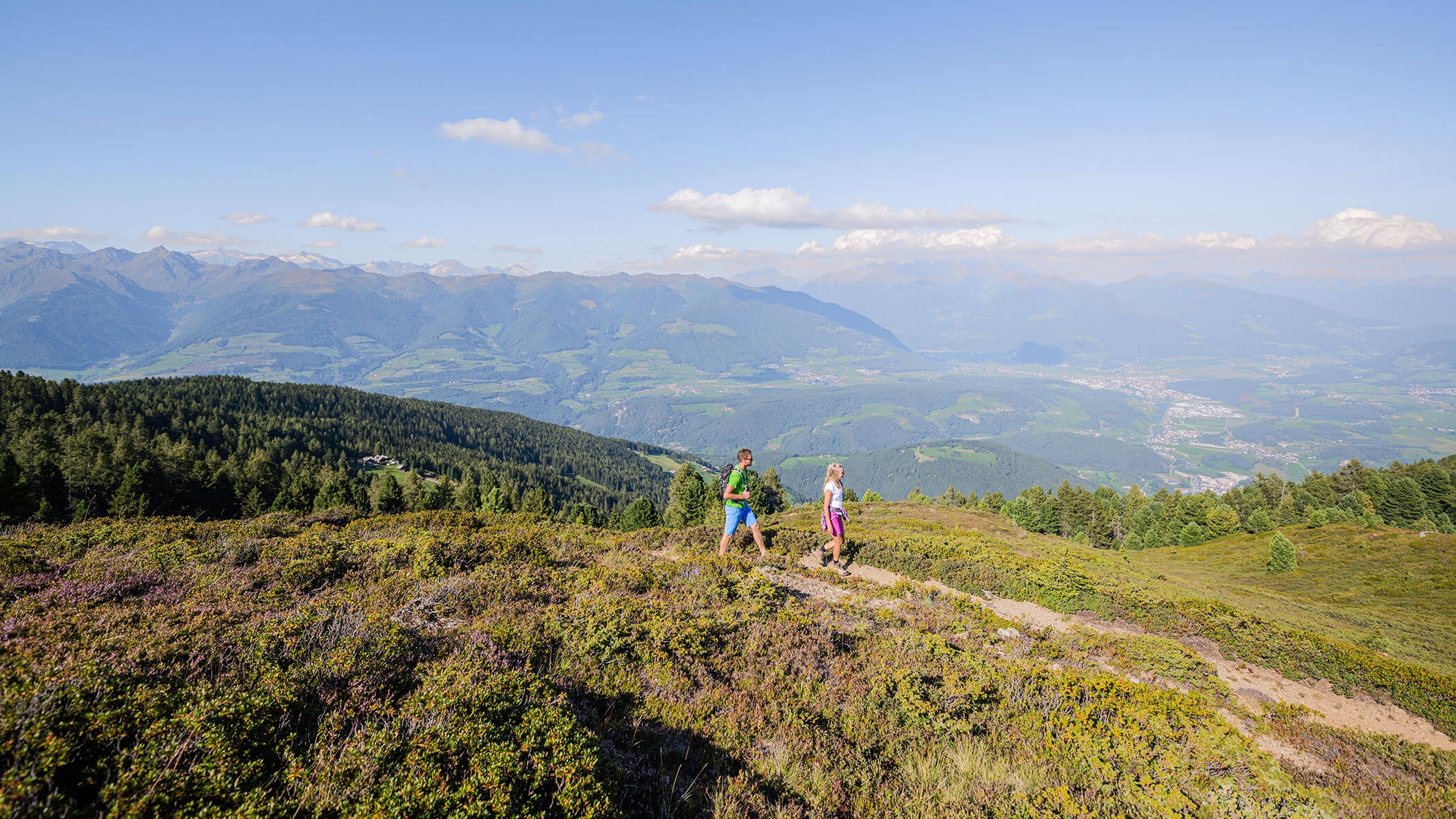 Zwei Menschen wandern auf einem grasbewachsenen Bergpfad mit malerischem Blick auf Wälder und ferne Berge unter blauem Himmel. - Lantl Deluxe Apartments
