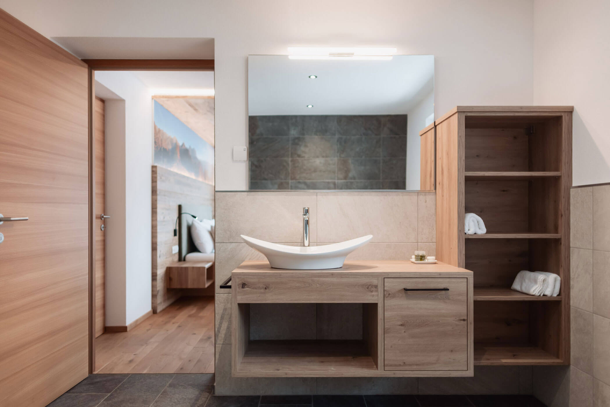 Modernes Badezimmer mit Waschtisch aus Holz, Aufsatzwaschbecken, großem Spiegel, offenen Regalen und Blick in ein Schlafzimmer. - Lantl Deluxe Apartments