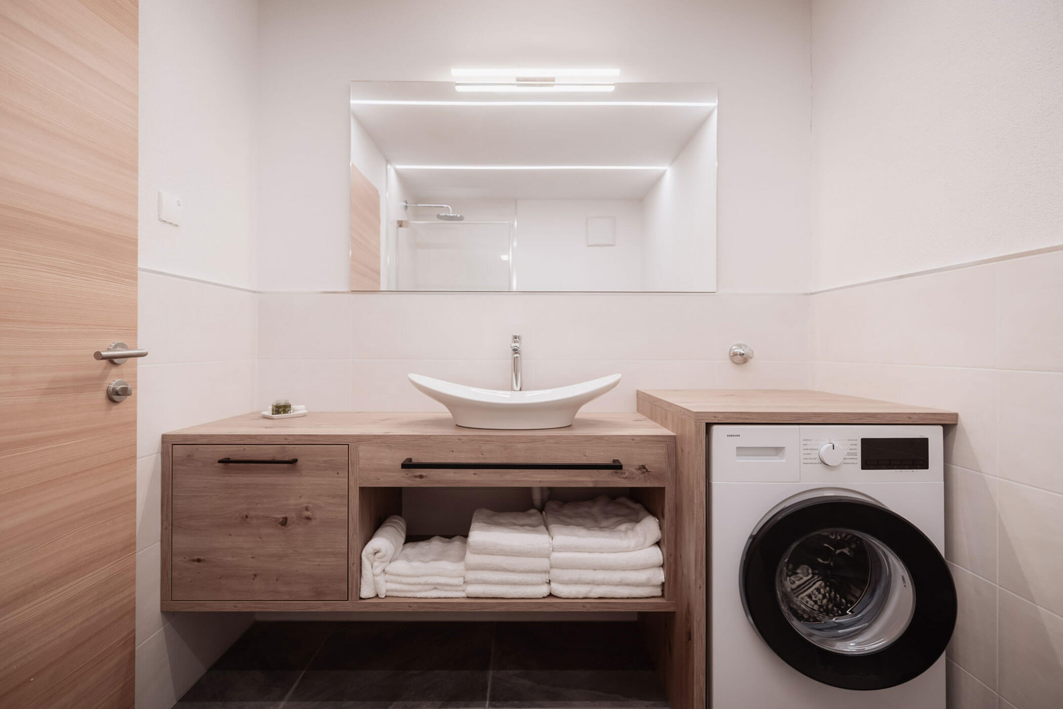 Modernes Badezimmer mit einem Waschtisch aus Holz, einem Aufsatzwaschbecken, Handtüchern und einer Waschmaschine unter einem großen Spiegel. - Lantl Deluxe Apartments