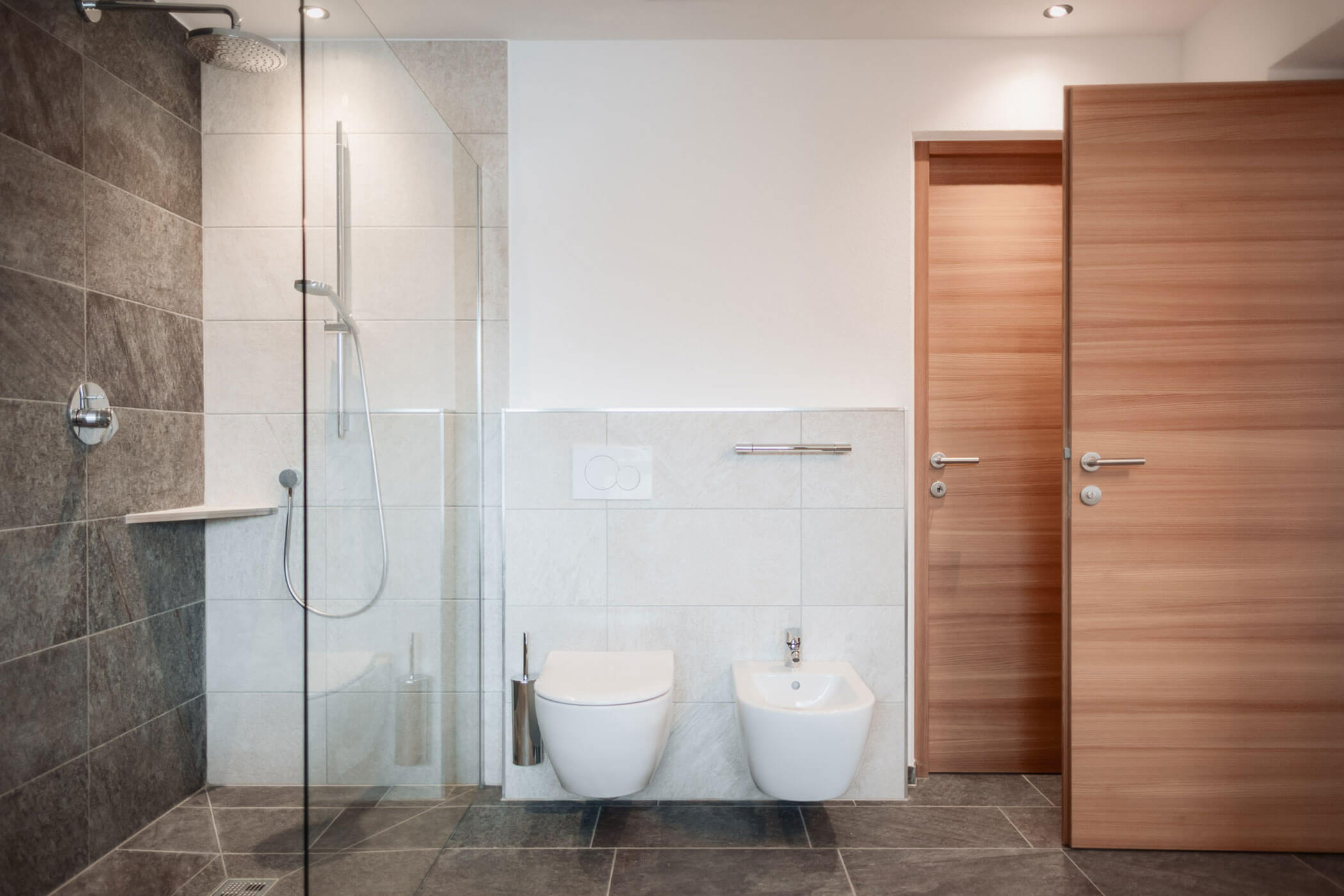 Modernes Badezimmer mit Glasduschkabine, wandhängendem WC, Bidet und Holztür auf einem mit Steinfliesen ausgelegten Boden. - Lantl Deluxe Apartments