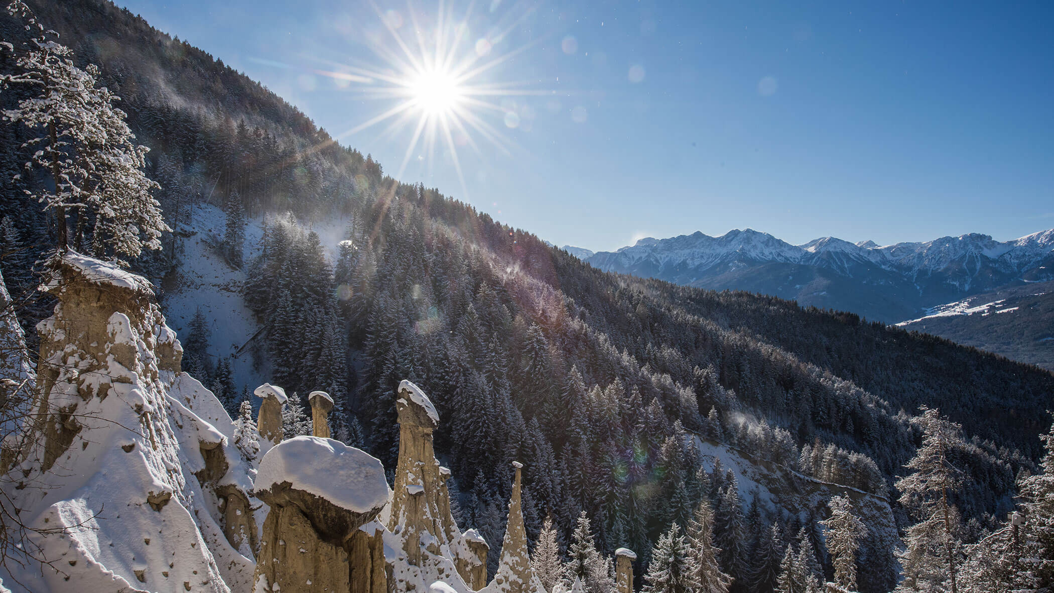 Schneebedeckte Felsformationen und bewaldete Berge unter strahlendem Sonnenschein und klarem blauem Himmel. - Lantl Deluxe Apartments