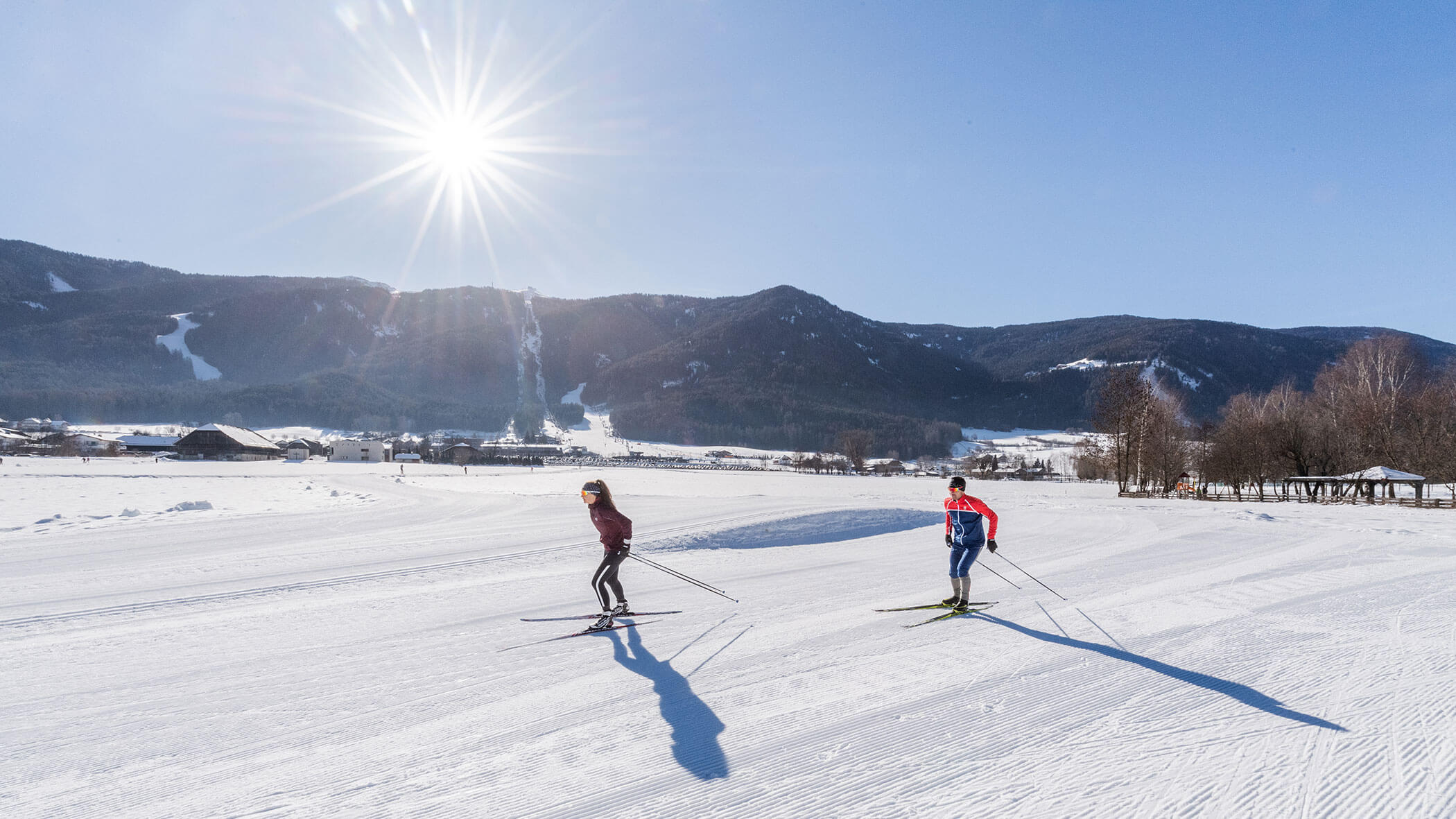Zwei Menschen fahren auf einem verschneiten Feld Langlauf, im Hintergrund sind Berge und ein strahlender Sonnenschein zu sehen. - Lantl Deluxe Apartments